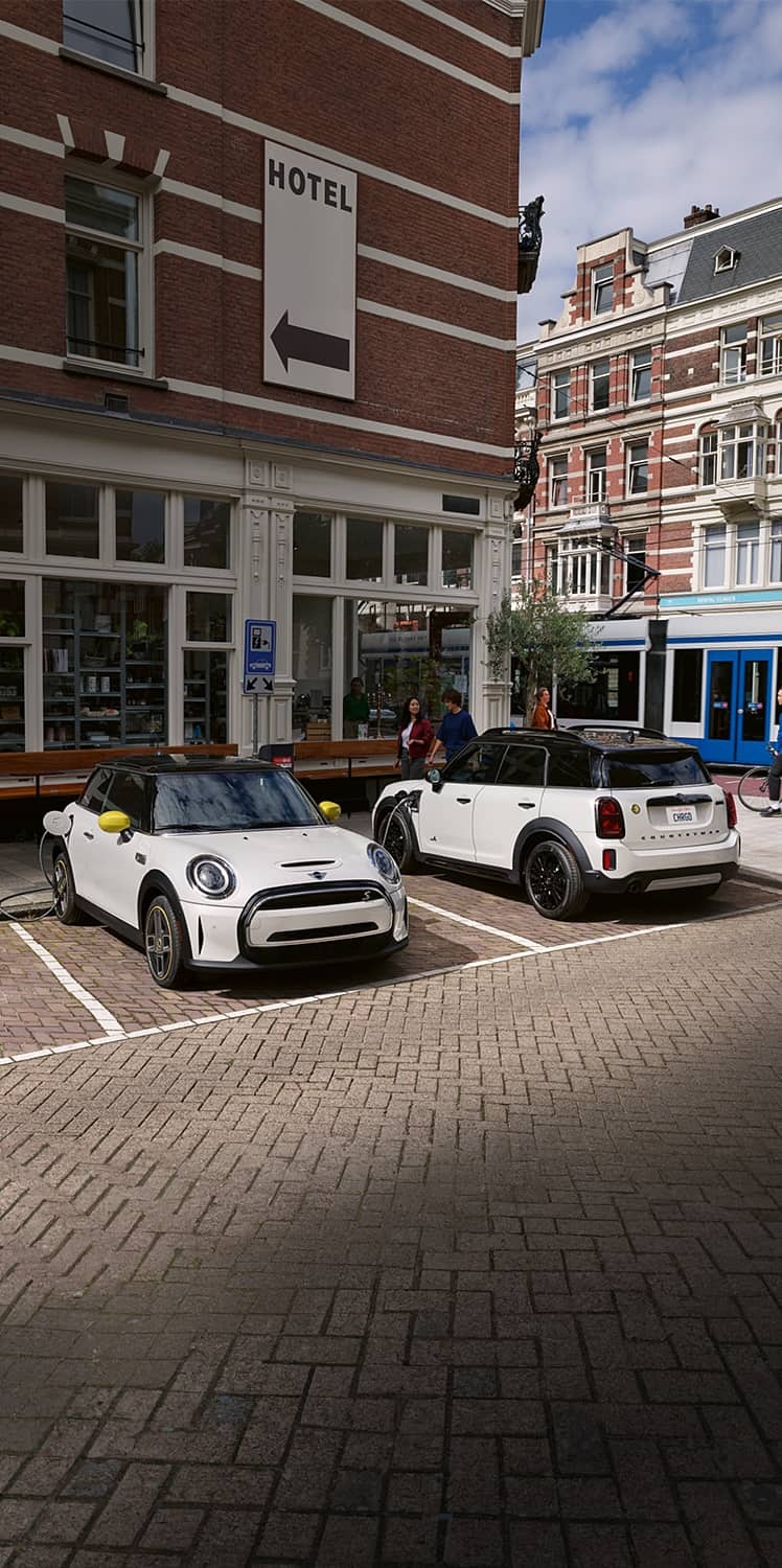Two MINI Electric vehicles parked in parking spots on a brick surface with retail storefronts and an urban setting in the background.