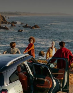 Three-quarters back view of a grey MINI vehicle with its doors open and people standing around it along with a wavy sea in the background.