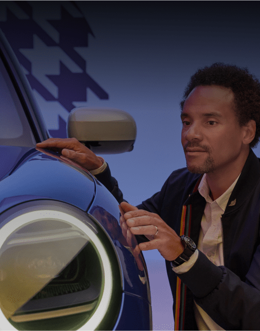 A man bending down to get a closer look at the headlight of a MINI against a purple background with a houndstooth pattern.