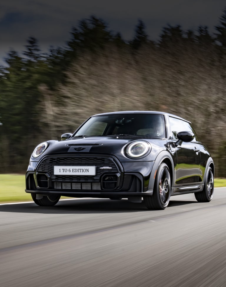 Three-quarters front view of a MINI John Cooper Works 1 To 6 Edition driving on a paved surface with a forest setting in the background along with clouds in the sky.