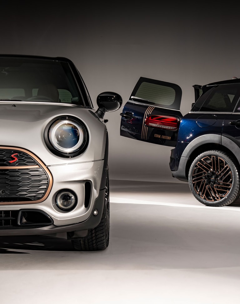 Front view of a MINI Clubman Final Edition in Nanuq White alongside another MINI Clubman Final Edition in Enigmatic Black, both of which are parked on a blank white surface with shadows underneath them and nothing in the background.