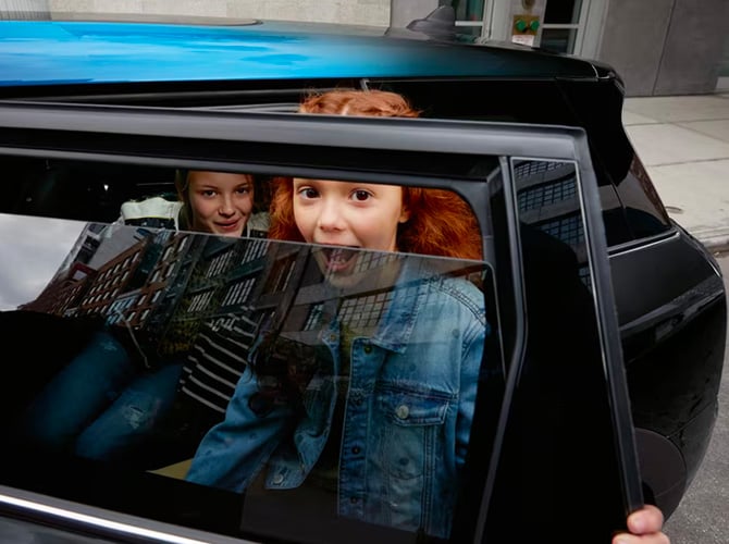 Closeup view of a blue MINI parked on a street surface with its back-left door open and two young girls grinning who are visible through the corresponding window that’s slightly open.