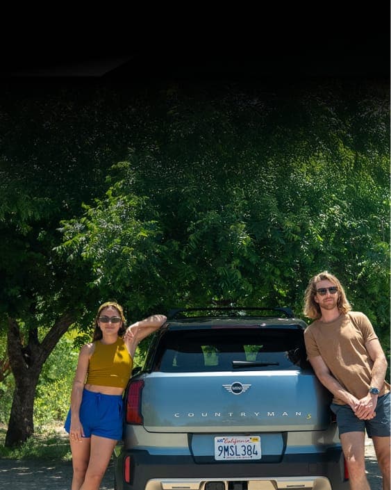 Lauren Bush and company lean on trunk of a MINI Countryman SE ALL4 with lush foliage in the background. 