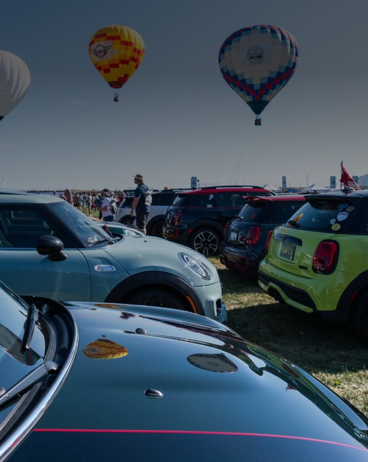 Side-view of rows of MINIs parked in a field during MINI Takes the States 2016 with hot air balloons in the distance.