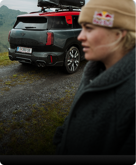 alt="Side profile portrait of Vali Höll and an angled rear view of the new MINI JCW Countryman ALL4, which we see equipped with a roof rack."