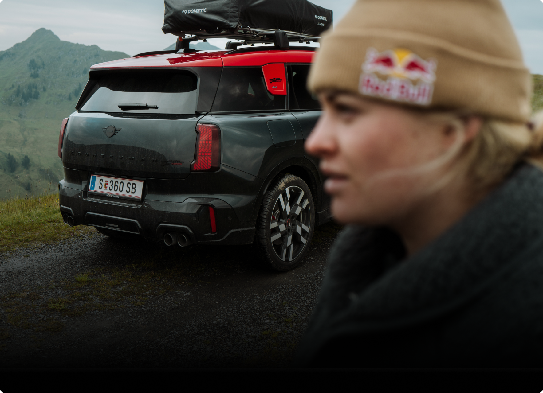 alt="Side profile portrait of Vali Höll and an angled rear view of the new MINI JCW Countryman ALL4, which we see equipped with a roof rack."