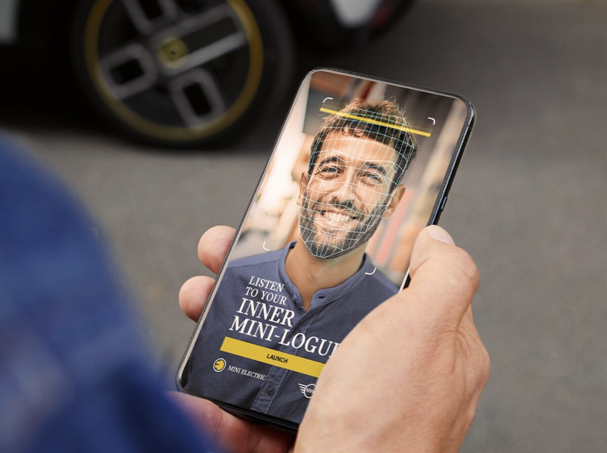 Light-skinned hand holding a smartphone with the MINI-logue AI experience displayed on screen, along with pavement blurred out in the background.