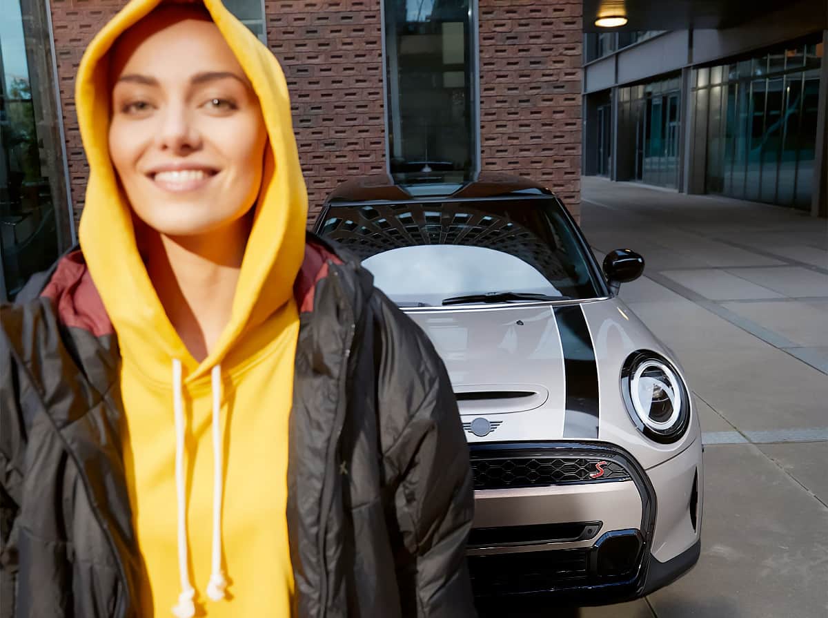 Grinning person wearing a yellow sweatshirt and black jacket, standing in front of a white MINI vehicle with black bonnet stripes and a brick building in the background.