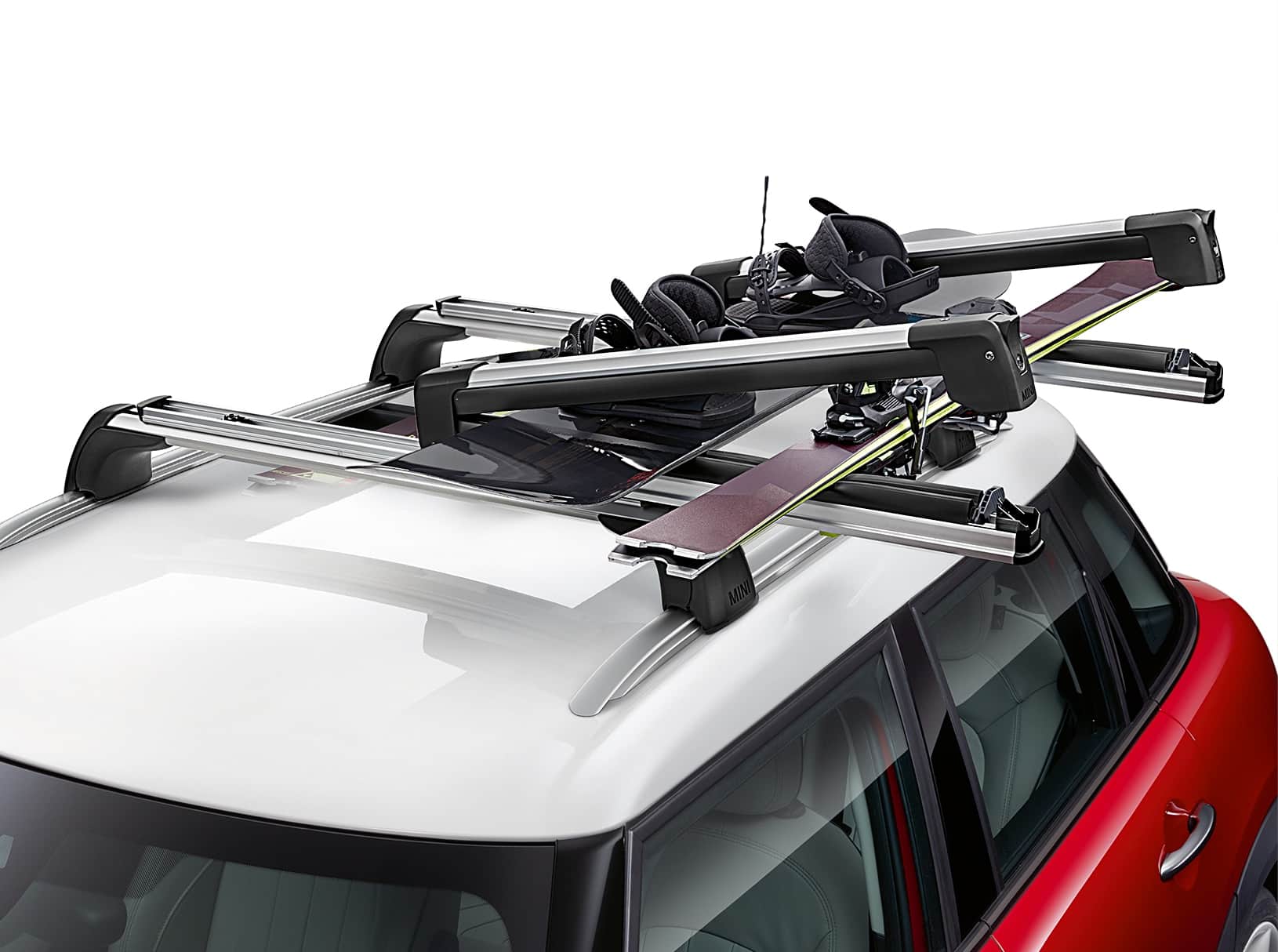 Closeup view of a white roof on a red MINI vehicle with a metal cargo rack on top of it, including a black snowboard with foot bindings and set of purple skis both fastened to the rack, in front of a blank white background.