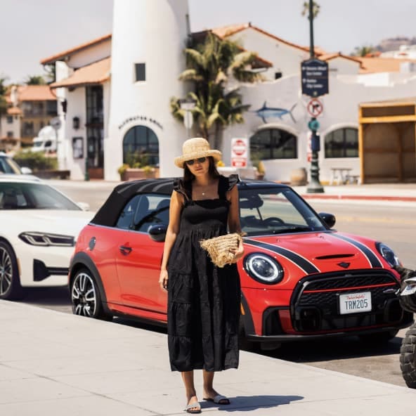 Woman in black dress and straw hat carrying a straw basket with a three-quarters front view of a red MINI with black bonnet stripes in the background parked on a street.