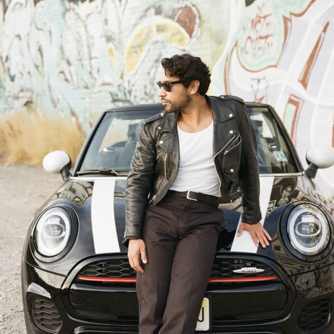 Man in a black coat, white shirt, and sunglasses standing in front of a black MINI with white bonnet stripes parked on a gravel surface, along with a wall containing graffiti in the background.