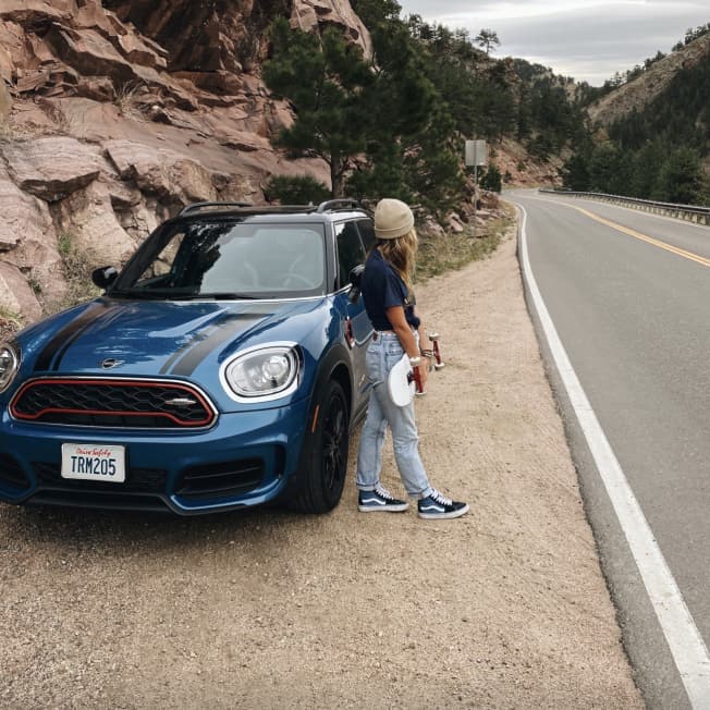 Front view of a blue MINI with black bonnet stripes parked on a gravel surface off to the side of an open road, with a woman wearing jeans and a knit beanie standing next to it.