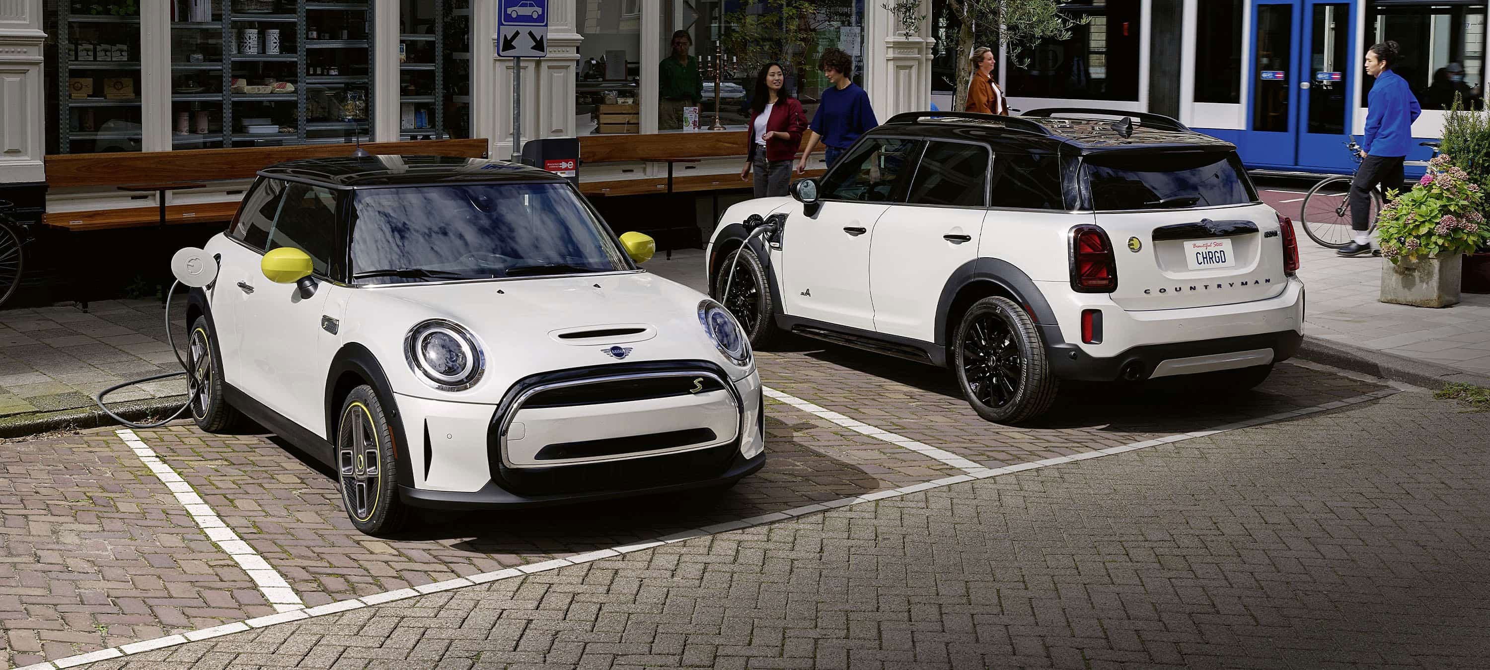 Two MINI Electric vehicles parked in parking spots on a brick surface with retail storefronts and an urban setting in the background.