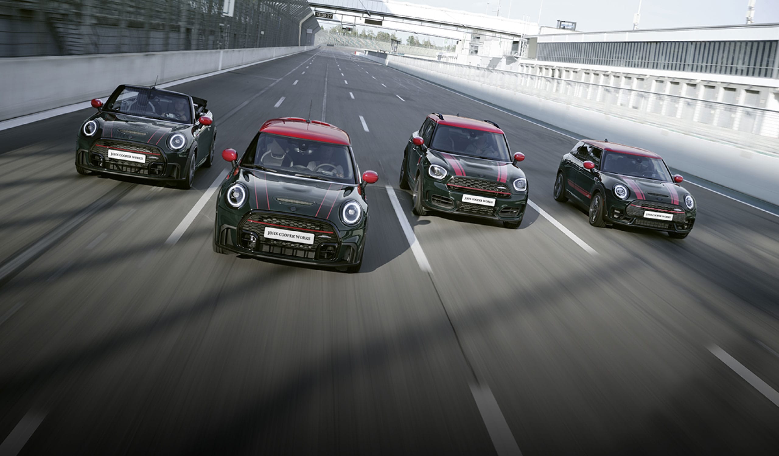 Front view of four dark colored MINI John Cooper Works vehicles racing alongside each other on a racetrack with white fencing on the right side.
