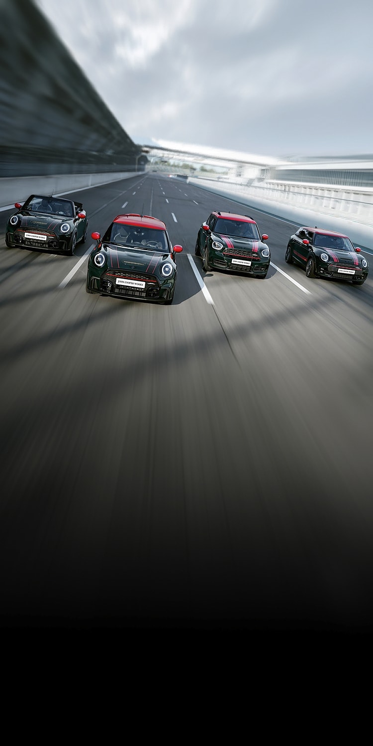 Front view of four dark colored MINI John Cooper Works vehicles racing alongside each other on a racetrack with white fencing on the right side.