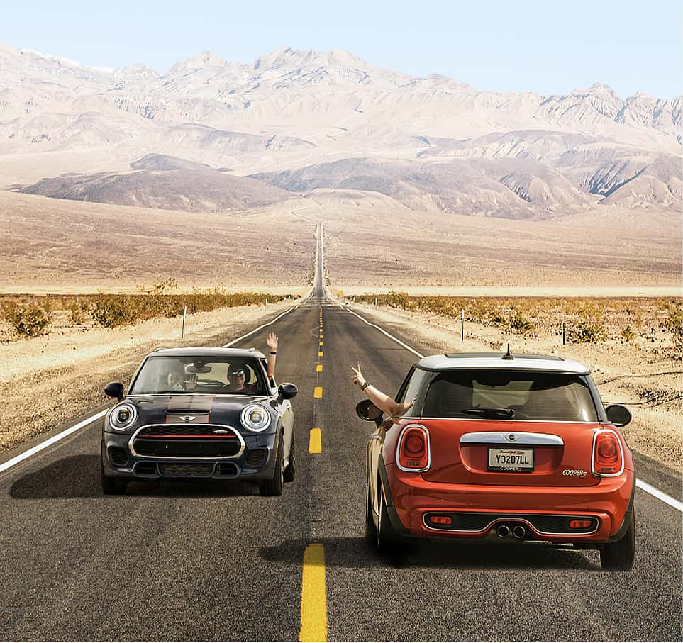 Two MINIs driving on an open road, one facing forward in black and one facing backwards in red, with mountains and clear skies in the background.