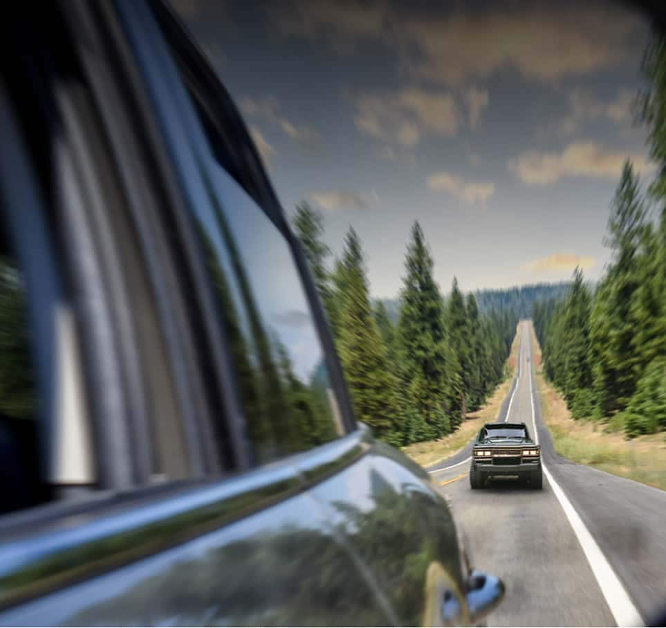 Fisheye view of a vehicle behind another vehicle driving on an open road with evergreen trees and clear skies in the background.