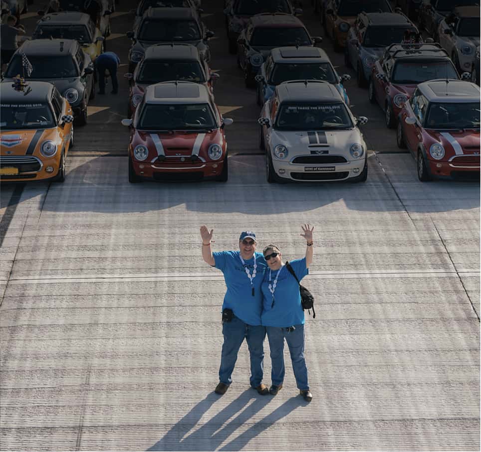 Overhead view of two people in blue shirts smiling and waving, with several MINI vehicles in different colors parked in a grid style in the background.