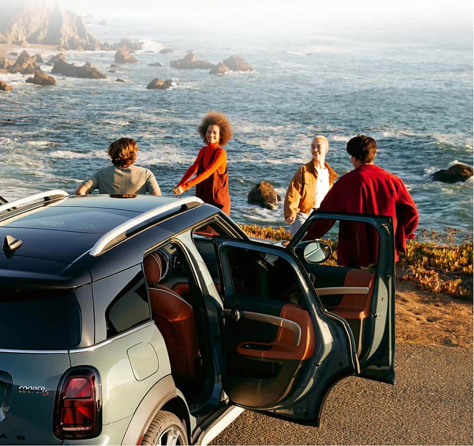 People standing around a MINI with its doors open with a body of water and mountain range in the background.
