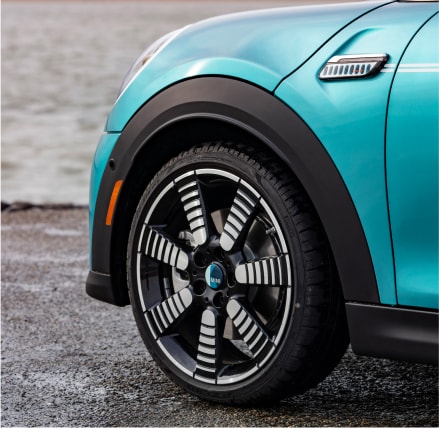 Closeup view of an 18” Pulse Spoke 2-Tone Wheel on a MINI Cooper S Convertible Seaside Edition in Caribbean Aqua, parked on a dark surface with a white wall in the background.