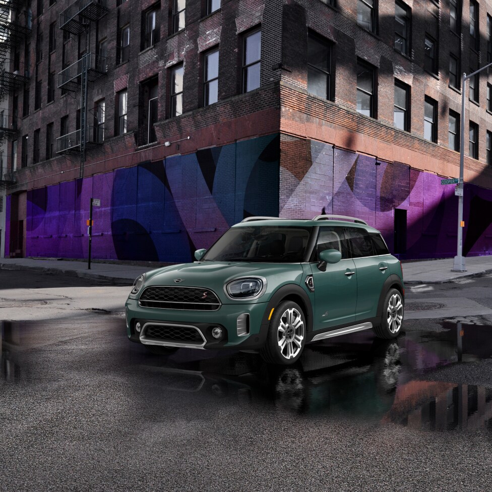 Three-quarters front view of a left-facing green 2024 MINI Cooper S Countryman parked on a street in an urban setting with puddles surrounding it and a brick building in the background.