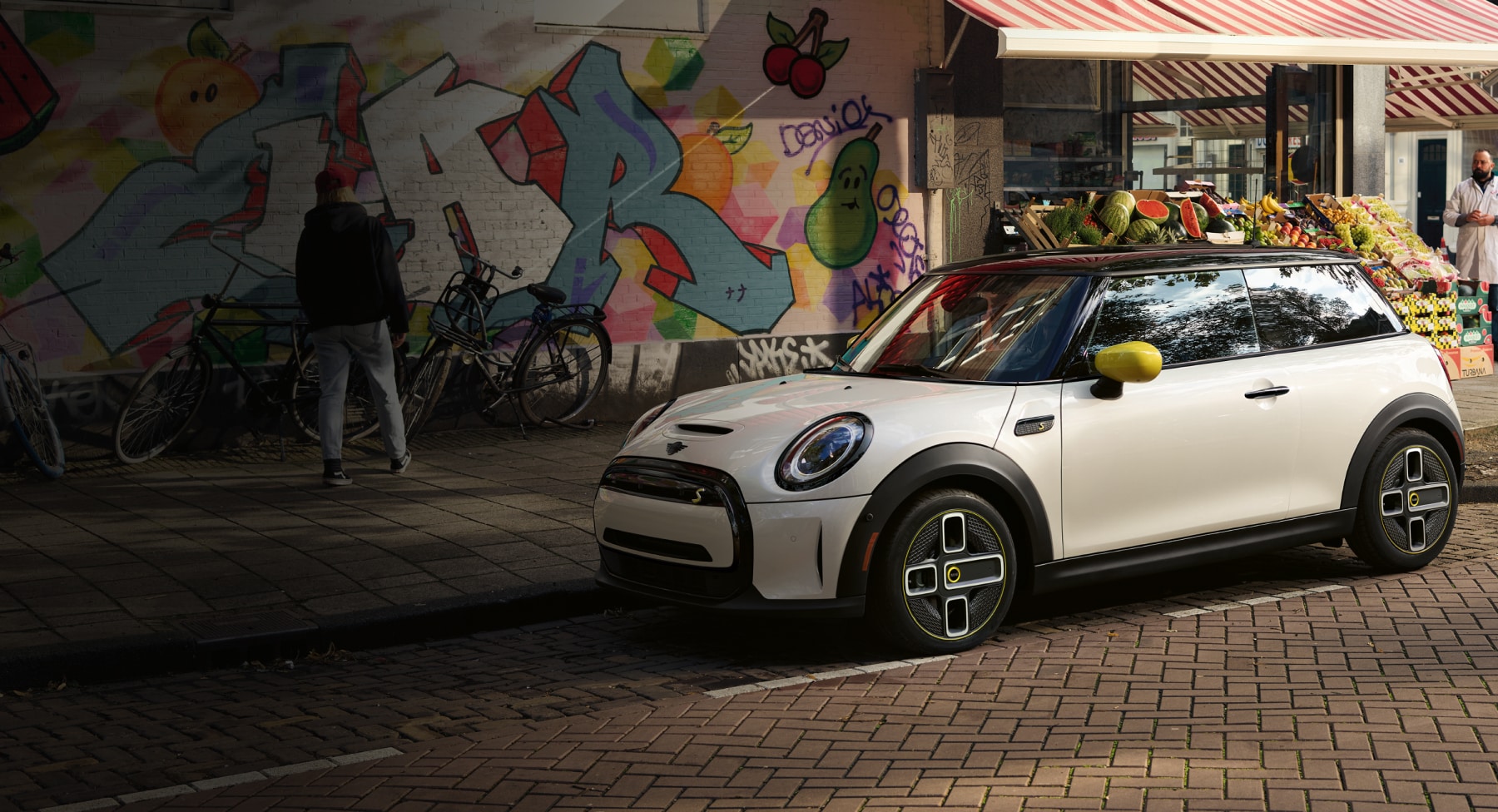 Three-quarters front view of a white MINI Cooper SE Hardtop 2 Door parked on a street in an urban setting with a graffiti wall and retail storefront in the background.