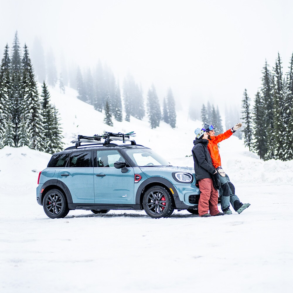 Side view of a light blue MINI vehicle parked in a mountainous setting with its shadow underneath it and two people in snow gear leaning into the hood of the vehicle, plus evergreen trees and snow falling in the background.