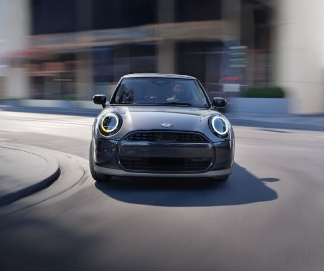 A MINI Cooper C 2 Door turns a corner in front of a modern looking building. The background has a motion blur on it.