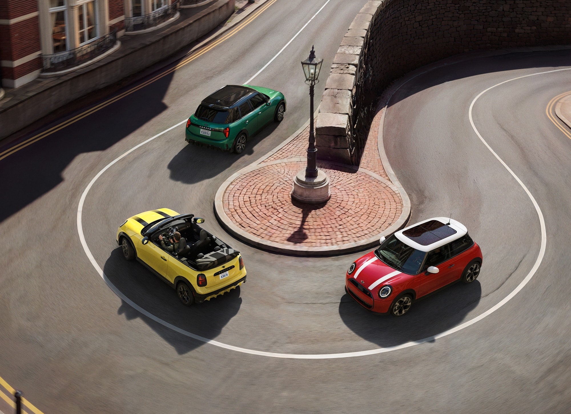 Bird’s-eye view of a MINI Cooper S 2 Door, MINI Cooper S Convertible, and MINI Cooper S 4 Door in different colors rounding a bend surrounded by charming city buildings. 