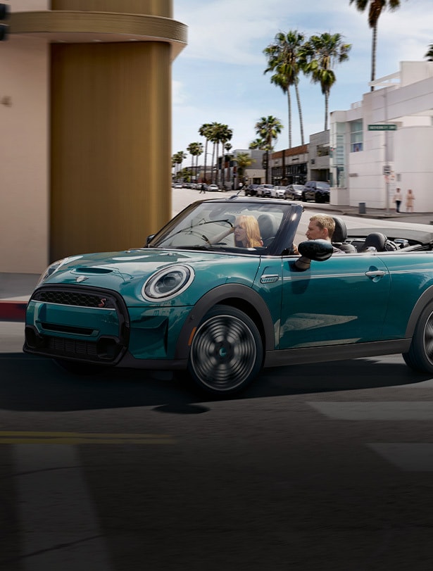 Three-quarters front view of a MINI Cooper S Convertible Seaside Edition in Caribbean Aqua rounding a street corner in an urban setting on a sunny day, with a man driving and a woman sitting in the front passenger’s seat.