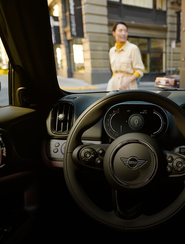 Driver’s seat view from the inside of a MINI vehicle, with a person in a pocketed off-white colored shirt and urban setting visible through the windshield.