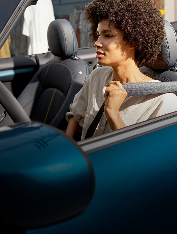 Woman in white shirt sitting in the driver’s seat of a blue MINI Convertible, holding the seatbelt with her left hand while looking onwards in the distance.