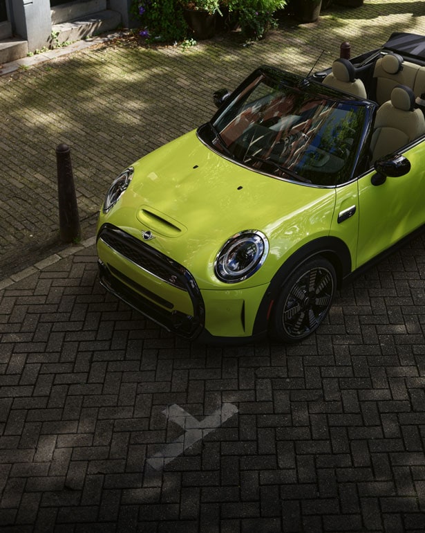 Three-quarters overhead view of a yellow MINI Convertible parked on a brick surface with a sidewalk in the background.